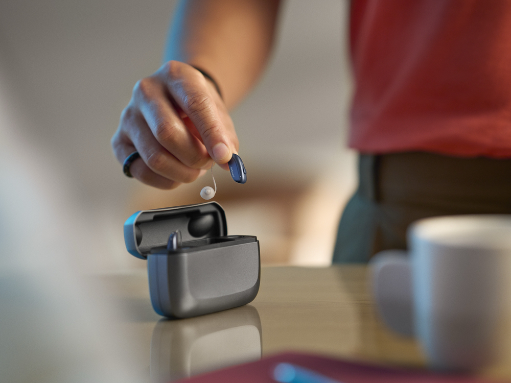 Patient lifting a rechargeable receiver-in-canal hearing aid from its charging case, a style fitted at AFA Balance & Hearing Institute in Mesa, AZ